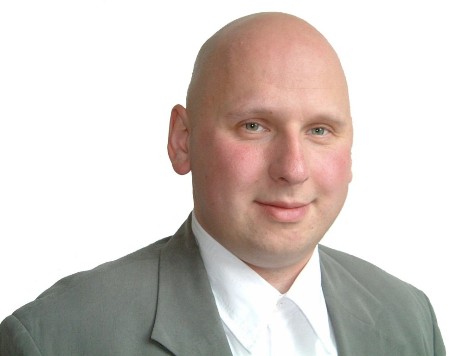 Ein glatzköpfiger Mann in einem grauen Anzug und einem weißen Hemd lächelt in die Kamera vor einem weißen Hintergrund.